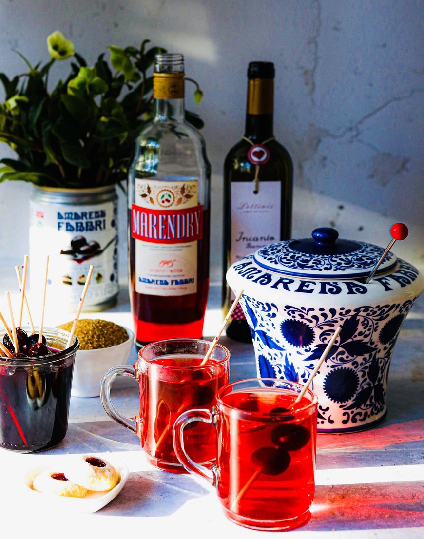 Amarena Affair, zwei Gläser roter Glühwein mit aufgespießten Amarenakirschen, dahinter eine Flasche Marendry, ein Weißwein und ein blau-weißes Amarena-Fabbri-Gefäß