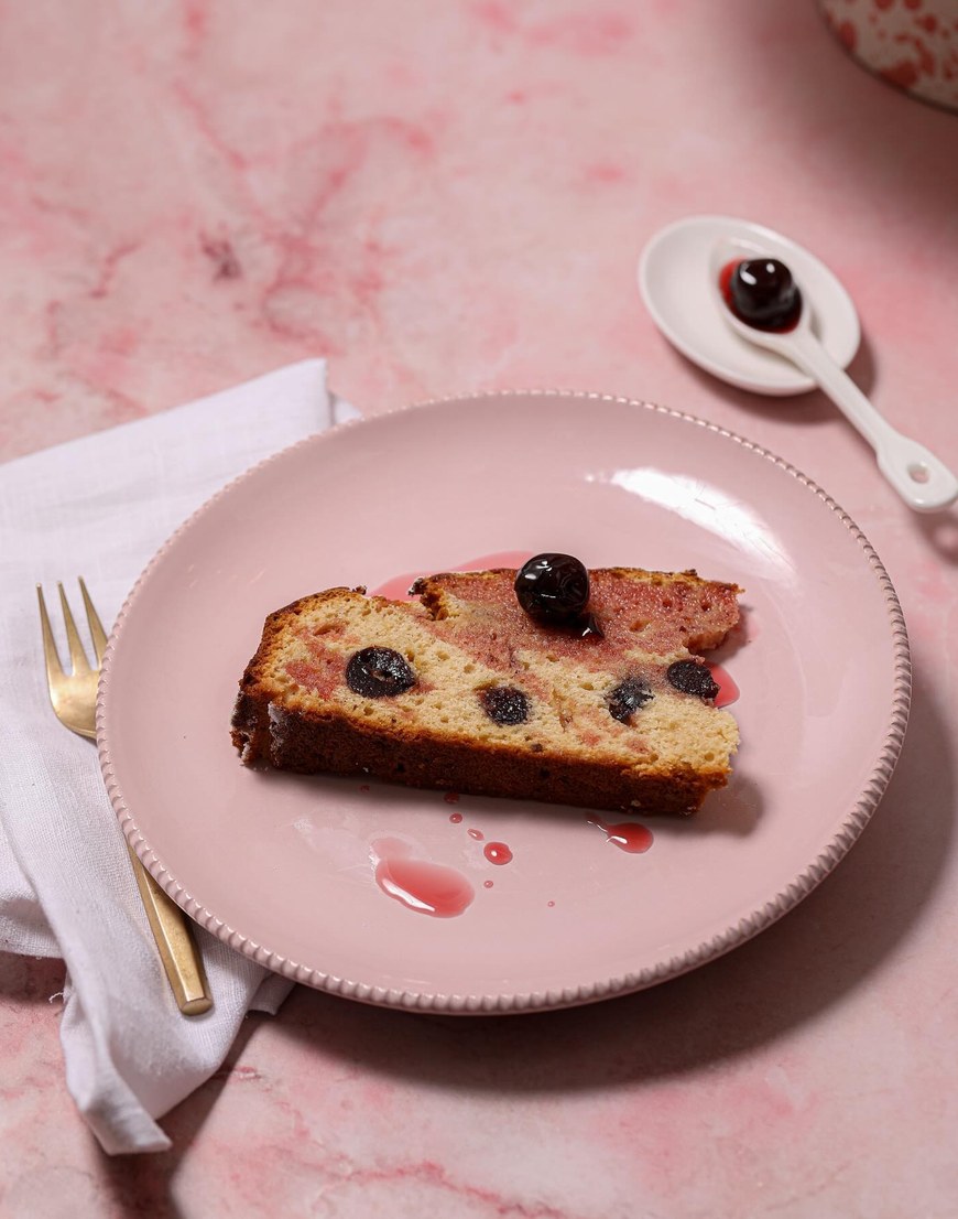 Dolce all’Amarena, ein Stück Amarena-Kuchen mit Kirschen und rosa Sirupschlieren auf einem rosa Teller, daneben eine Amarenakirsche auf einem Porzellanlöffel