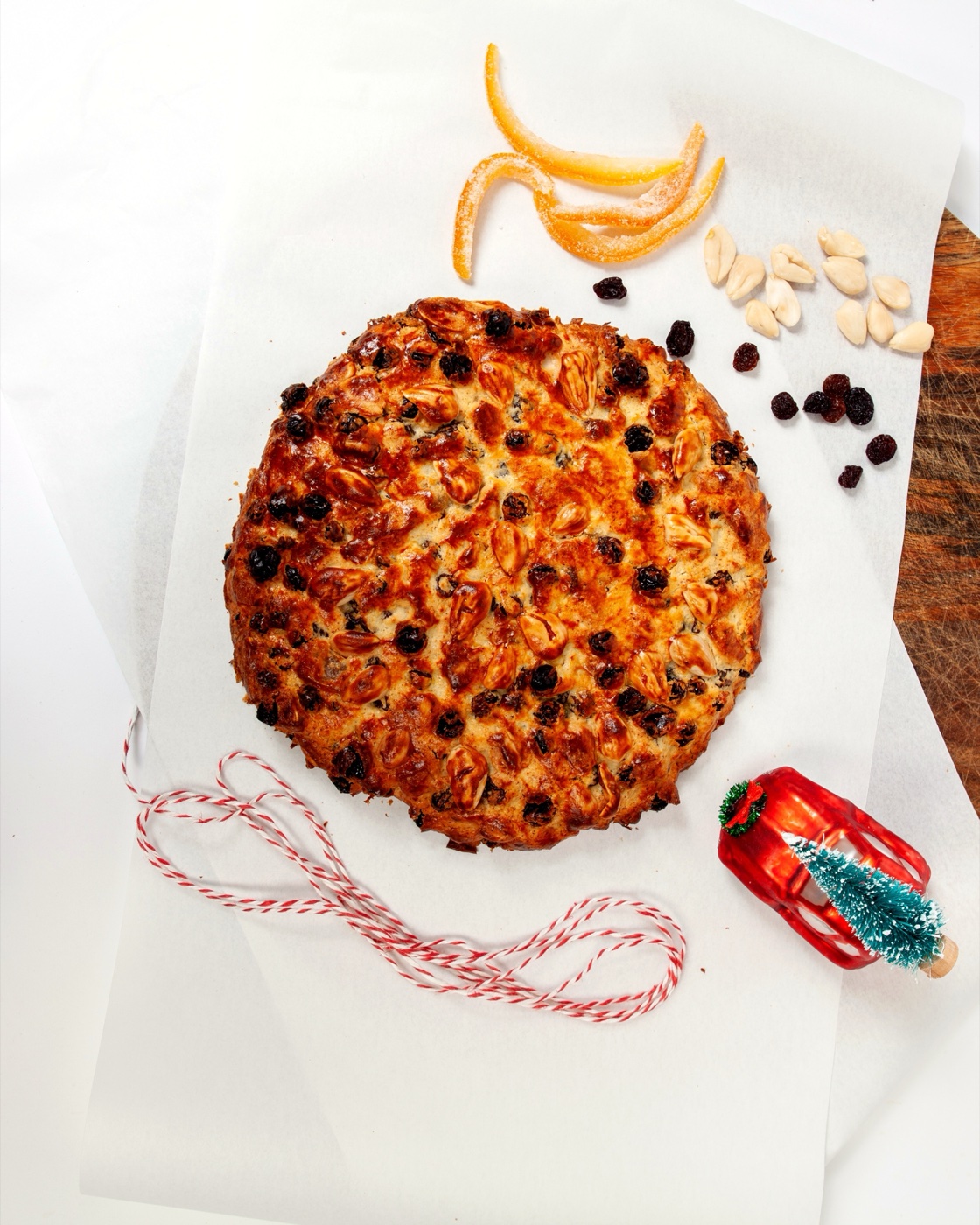 Flacher Pandolce Genovese mit Mandeln und Rosinen auf Backpapier, daneben kandierte Orangenstreifen, Mandeln, eine rot-weiße Kordel und ein kleines rotes Spielzeugauto mit Tannenbaum