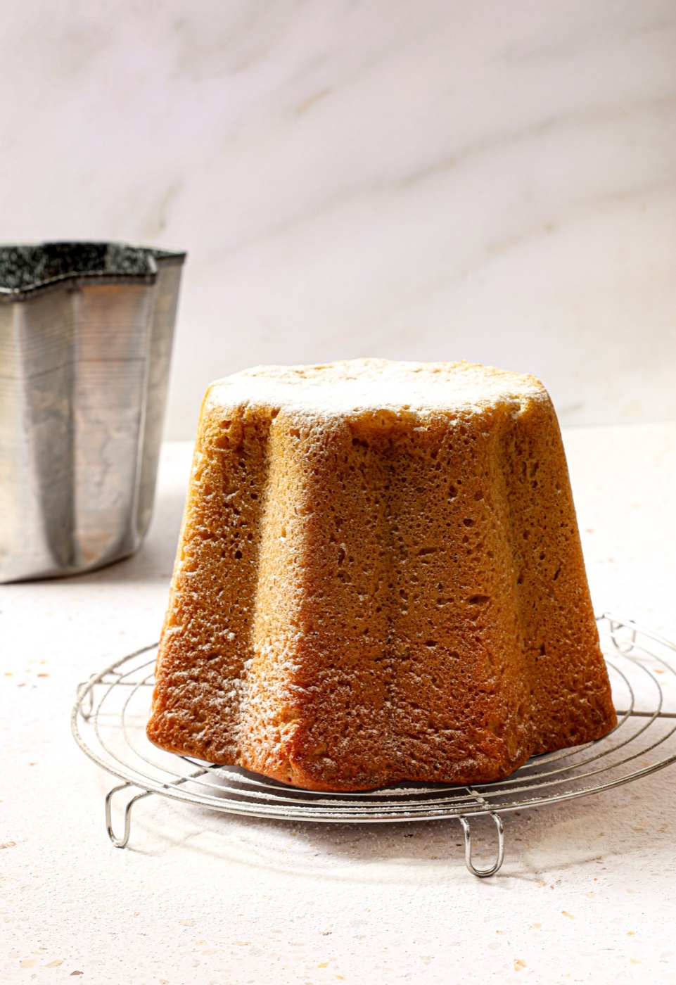 Selbst gebackener Pandoro in Sternform mit Puderzucker auf einem Kuchengitter, dahinter die leere Pandoro-Form vor heller Marmorwand