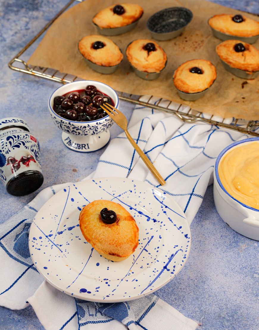 Pasticciotti mit Amarena, goldbraune ovale Mürbeteigtörtchen mit je einer Amarenakirsche obenauf, eines auf einem blau gesprenkelten Teller, dahinter Kirschen in einer Fabbri-Schale und Vanillecreme